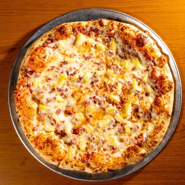 A whole Hawaiian pizza with pineapple and ham on a metal pan, on a wooden table.