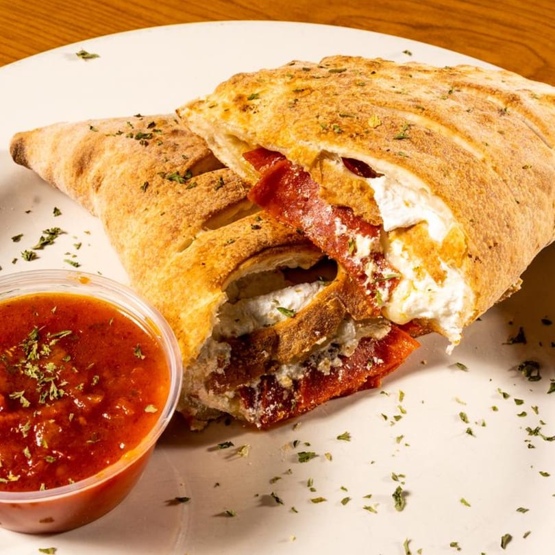 A golden-brown calzone, sliced open to reveal ricotta cheese and pepperoni, served with marinara sauce.